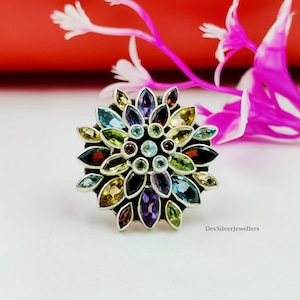 Puede incluir: Un anillo de plata con un diseño floral, con una variedad de piedras preciosas de colores. Los pétalos del anillo están adornados con piedras azules, verdes, amarillas, moradas y rojas. El anillo se encuentra sobre un fondo borroso con un elemento floral rojo y rosa.