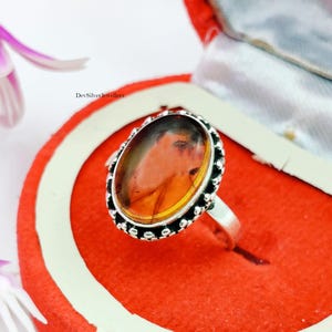 Könnte beinhalten: Ovaler Bernsteinring mit Silberband. Der Bernsteinstein hat eine warme, orange-braune Farbe und ist in einer dekorativen Silberfassung gefasst. Der Ring wird in einer roten Schmuckschatulle präsentiert. Der Text "DevSilverJewellers" ist sichtbar.