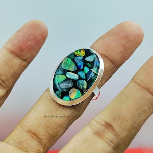 Könnte beinhalten: Ovaler Silberring mit einem Mosaik aus farbigen, unregelmäßig geformten Steinen auf schwarzem Hintergrund. Die Steine zeigen Grün-, Blau-, Gelb- und schillernde Farbtöne. Der Ring wird an einem Finger getragen.