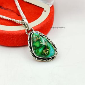 Puede incluir: Un collar con colgante de plata con una piedra verde y turquesa en forma de lágrima. La piedra está engastada en un marco de plata con un borde en forma de cuerda. El collar se exhibe sobre una superficie roja.