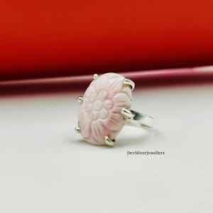Può includere: Un anello rosa chiaro con un design floreale intagliato. La gemma ovale è incastonata in una fascia d'argento con quattro griffe. L'anello è esposto su una superficie bianca con sfondo rosso. Il testo "DevSilverJewellers" è visibile.
