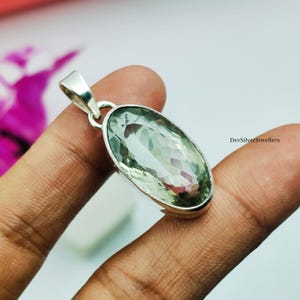 Puede incluir: Un colgante de plata con una piedra preciosa ovalada de color verde claro. La piedra preciosa tiene un aspecto sutil y multicolor. El colgante tiene un engaste sencillo de plata pulida y un pequeño lazo para una cadena. El fondo es una mezcla borrosa de colores.