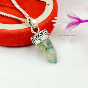 Puede incluir: Collar con colgante de plata con un cristal facetado verde claro con inclusiones oscuras. El cristal está engastado en una montura de plata decorativa y cuelga de una cadena de plata. El fondo incluye una superficie roja y una flor.