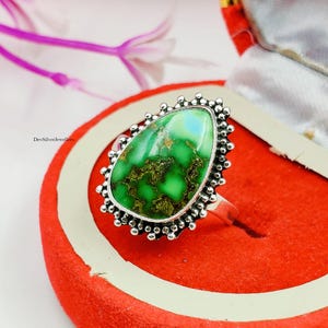 Puede incluir: Un anillo de plata con una piedra verde grande en forma de lágrima. La piedra tiene un patrón moteado de verde y marrón, con un área azul claro. El anillo está engastado en un bisel de plata con un borde de cuentas.
