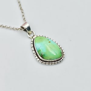 Può includere: Una collana con pendente a forma di goccia caratterizzata da una pietra liscia verde chiaro con sottili inclusioni scure. La pietra è incastonata in una cornice d'argento lucido con bordo dentellato, sospesa a una sottile catena d'argento.