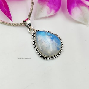 Puede incluir: Un collar con colgante de plata con una piedra lunar en forma de pera. La piedra iridiscente está engastada en un marco de plata decorativo y cuelga de una cadena de plata trenzada. El fondo incluye acentos florales rosas y blancos.