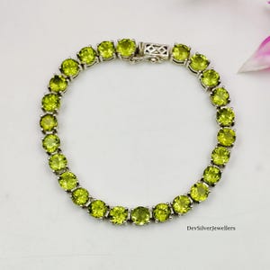 Puede incluir: Una pulsera de plata con una serie de piedras preciosas redondas de color verde claro. Las piedras están engastadas muy juntas, creando una línea continua de color. La pulsera tiene un cierre seguro y se muestra sobre una superficie blanca.