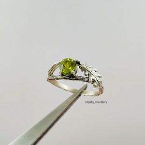 Op de afbeelding: Een zilveren ring met een lichtgroene, ronde edelsteen. De ring heeft een op de natuur geïnspireerd ontwerp met een takachtige band en een blad. De tekst "Dipikajewellers" is zichtbaar.