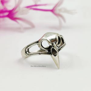 Könnte beinhalten: Ein silberner Ring mit einem Vogelschädel-Motiv. Der Ring zeigt einen detaillierten, dreidimensionalen Schädel mit Augenhöhlen und einem spitzen Schnabel. Das Band hat ein offenes Design. Der Text "DevSilverJewellers" ist auf dem Ring sichtbar.
