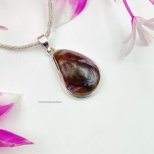 Puede incluir: Colgante plateado con una piedra marrón y púrpura en forma de lágrima. El colgante cuelga de una cadena plateada. En el fondo se ven pétalos de flores rosas y blancas.