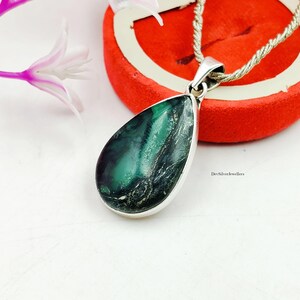 Puede incluir: Un colgante en forma de lágrima con un bisel plateado y una piedra verde y negra. El colgante cuelga de una cadena plateada. El fondo incluye una caja roja y una superficie blanca.