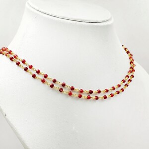 May include: A double-strand necklace with small, round, red beads strung on delicate gold-coloured chains. The necklace is displayed on a white mannequin, highlighting its design and colour contrast.