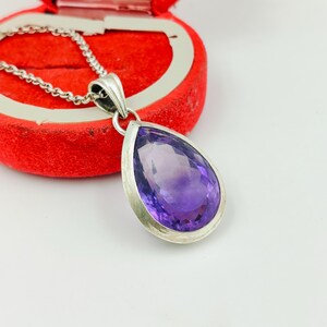 Könnte beinhalten: Ein tropfenförmiger Amethyst-Anhänger in einer silberfarbenen Metallfassung, der an einer passenden Gliederkette hängt. Der violette Edelstein hat eine facettierte Oberfläche, die das Licht reflektiert und klar glänzt.