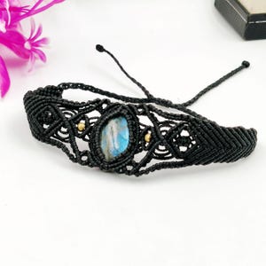 May include: A black macramé bracelet with a central oval, blue-grey gemstone. The bracelet has a woven design with small gold-coloured beads and adjustable ties. The jewellery is displayed on a white surface.