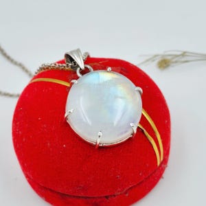 Op de afbeelding: Een ronde, in cabochon geslepen maansteen hanger in een zilverkleurige metalen zetting met klauwen. De edelsteen heeft een doorschijnend, melkachtig wit uiterlijk met subtiele iriserende blauwe en gele flitsen. Een dunne zilverkleurige ketting is bevestigd.