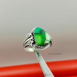 Puede incluir: Un anillo de plata con una piedra grande en forma de pera, verde y blanca. El anillo tiene un diseño de filigrana ornamentado en la banda y alrededor de la piedra. El anillo está sujeto por unas pinzas. El patrón de la piedra es verde y blanco.