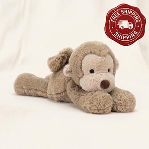 May include: A plush monkey toy made of soft, light brown fur. The stuffed animal is in a lying position with its front paws extended forward. It features a lighter beige face, small rounded ears, black eyes, and a dark brown nose.