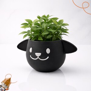 Peut inclure: Un pot de fleurs en céramique noire en forme de tête de mouton, avec des traits blancs et des oreilles. Le pot est rempli de plantes vertes luxuriantes, créant un élément décoratif charmant.