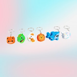 May include: A collection of colorful animal-shaped keychains. The keychains include a cat, dinosaur, dog, lion, shark, and unicorn. Each keychain is made of a soft material and has a silver metal ring for attaching keys.