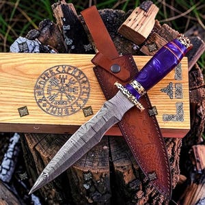 May include: A Damascus steel dagger with a purple handle and gold accents, resting on a wooden box with a leather sheath. The box features a circular design and the dagger is displayed on a bed of wood.