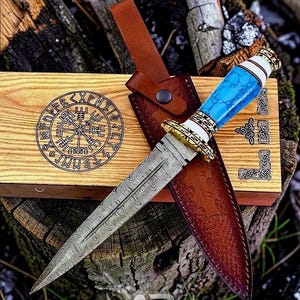 Puede incluir: Un cuchillo de acero de Damasco con mango azul y funda de cuero, presentado en una caja de madera con un diseño de rosa de los vientos. El cuchillo tiene una guarnición dorada decorativa y una base de mango blanca. La caja y la funda son marrones.