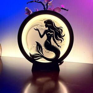 May include: A circular black lamp featuring a backlit silhouette of a mermaid with long flowing hair. The mermaid is positioned in the center against a glowing white background with stylized wave patterns. The lamp has a sturdy base and emits light.