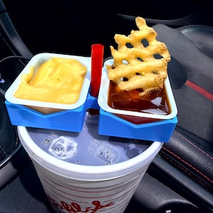 May include: A fast food meal featuring a cup of soda, a blue 3D-printed holder with two sauce containers, and a red straw. One container holds cheese sauce, the other holds dipping sauce, and a waffle fry sits atop the sauce.