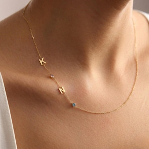 May include: A delicate gold necklace featuring the letters "K" and "N" with a pink and blue gemstone. The necklace is a fine chain, and the letters are small and elegant. The necklace is worn around the neck.
