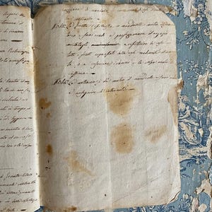 May include: An antique, off-white paper document featuring handwritten cursive text in black ink. The paper shows signs of age with visible brown stains and frayed edges. The document is positioned against a blue and white floral patterned surface.