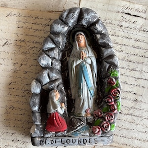 Può includere: Una statuetta modellata che raffigura una figura in veste bianca con fascia blu all'interno di una grotta in pietra grigia, con una figura più piccola inginocchiata con gonna rossa e velo bianco, rose rosse e la scritta M. DI LOURDES alla base.