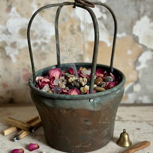 Può includere: Un pentolino in rame usurato con manico in metallo, riempito di petali di rosa essiccati, piccoli sassi e cristalli. Un sottile filo di fumo sale dal centro. Vicino si trovano bastoncini di legno, un piccolo campanello in ottone e un cucchiaio.