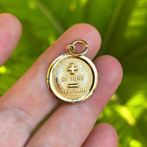 Può includere: Un ciondolo rotondo in oro con un simbolo a croce centrale sopra il testo DI IERI e DI DOMANI, caratterizzato da un motivo a raggiera che si irradia dal centro. Il ciondolo include un piccolo anello circolare fissato in alto.
