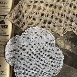 Può includere: Due centrini fatti a mano all'uncinetto. Uno è di forma rettangolare beige chiaro con un bordo smerlato. L'altro è di forma bianca a quattro lobi. Entrambi sono posizionati sopra un vecchio giornale aperto.