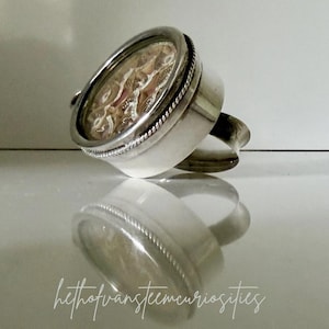 Pode incluir: Um anel de prata com uma face oval e texturizada. O anel tem uma borda decorativa em forma de corda e uma faixa larga. A face do anel tem um design detalhado em relevo. O anel é exibido em uma superfície reflexiva.