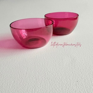 Può includere: Due piccole ciotole rotonde in vetro magenta traslucido con bordi lisci e curvi. Le ciotole sono posizionate una leggermente dietro l'altra, mostrando una tonalità rosa vibrante e un design semplice e aperto. Il testo hethofvansteencuriosities è visibile.
