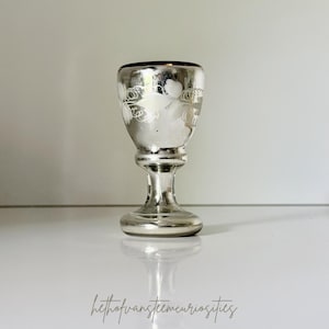 Op de afbeelding: Een zilverkleurig glas in vintage-stijl met een decoratief bloemenpatroon. Het glas heeft een brede kom, een slanke steel en een ronde voet. De rand van het glas heeft een donkere, reflecterende kleur. Het glas staat op een wit oppervlak.
