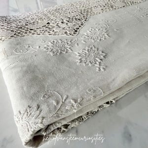 Pode incluir: Um têxtil de linho branco sujo dobrado com bordados florais intrincados e uma larga borda de renda decorativa. O tecido exibe padrões de costura detalhados, incluindo motivos de flores bordadas e desenhos de vinhas ao longo das bordas.
