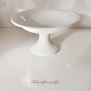 May include: A white ceramic pedestal bowl with a smooth, glossy finish, featuring a wide, shallow basin supported by a sturdy, flared base. The text "hethofvansteemcuriosities" is printed in a thin, brown, cursive font at the bottom center.