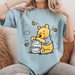 Puede incluir: Camiseta azul claro con una ilustración de dibujos animados de Winnie the Pooh, un oso amarillo con una camisa azul, con un tarro de miel etiquetado "Hunny" y abejas. El diseño está impreso en una camiseta azul claro. Las abejas son negras y amarillas.