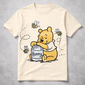 May include: Cream-colored t-shirt featuring a cartoon illustration of Winnie the Pooh with a honey pot labeled "Hunny." Bees fly around the bear. The shirt is a classic crew neck style.
