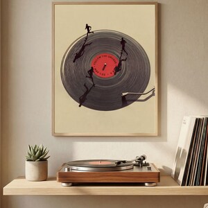 May include: A framed art print featuring a vinyl record with silhouetted figures running on its surface. The record has a red center with white text. A wooden record player sits below the print on a shelf, with a small potted plant and a stack of vinyl records.