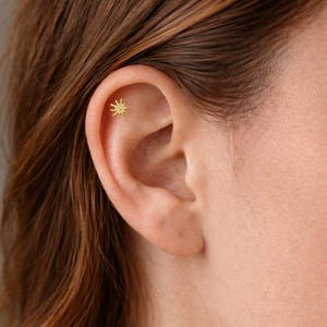 Può includere: Primo piano di un orecchio con un piccolo orecchino dorato a forma di sole. L'orecchino è a lobo, con un design dettagliato di raggi che emanano da un cerchio centrale. La persona ha la pelle chiara e i capelli castani.