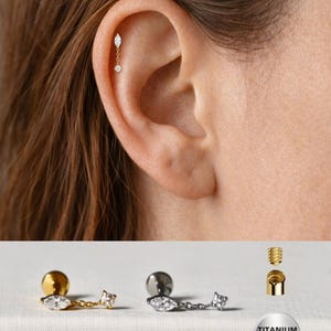Peut inclure: Boucle d'oreille hélix dorée avec une chaîne et deux pierres transparentes, portée à l'oreille. En dessous, une boucle d'oreille dorée et argentée est affichée, ainsi qu'un schéma des composants de la boucle d'oreille. Le mot "Titanium" est visible.