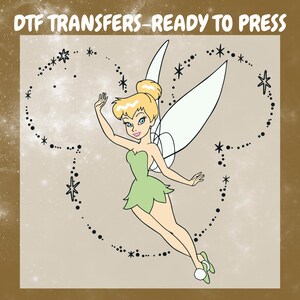May include: A DTF transfer ready to press featuring a cartoon fairy with blonde hair in a bun, wearing a green dress and white slippers. The fairy is surrounded by a dotted outline of a mouse head shape with star accents. Text reads DTF TRANSFERS-READY TO PRESS.