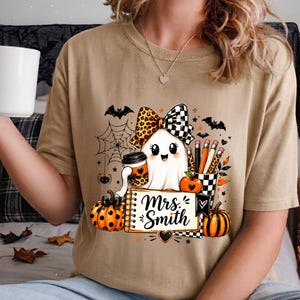 Może przedstawiać: Beżowy t-shirt z grafiką ducha w panterkę i w kratkę, trzymającego kubek kawy. Wz&oacute;r zawiera dynie, oł&oacute;wki, pajęczynę, nietoperze oraz notatnik z napisem Mrs. Smith wykonanym czarną czcionką przypominającą pismo odręczne.