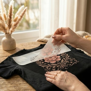 May include: A black t-shirt featuring a floral graphic design with pink and gold tones. A clear transfer sheet is being peeled back from the fabric, revealing the intricate botanical illustration of a large flower surrounded by ornate swirling vines.