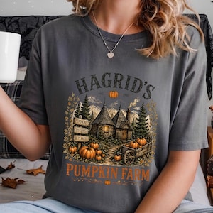 May include: A charcoal gray t-shirt featuring a graphic print of a rustic cottage surrounded by pine trees and pumpkins. The text reads HAGRID'S est. 1981 PUMPKIN FARM, with smaller signs labeled PUMPKINS, POTIONS, & PLANTS. The design includes a wooden cart.