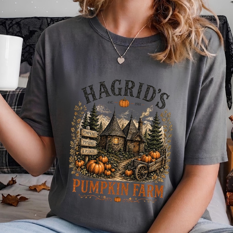 May include: A charcoal gray t-shirt featuring a graphic print of a rustic cottage surrounded by pine trees and pumpkins. The text reads HAGRID'S est. 1981 PUMPKIN FARM, with smaller signs labeled PUMPKINS, POTIONS, & PLANTS. The design includes a wooden cart.