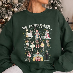 May include: A dark green crewneck sweatshirt featuring a graphic print titled THE NUTCRACKER. The design displays various characters from the ballet, including a nutcracker, Clara, the Mouse King, and dancers, accented with candy canes and stars.