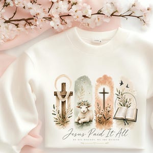 May include: A cream-colored crewneck sweatshirt featuring a graphic print of a wooden cross, a lamb with a floral crown, a second cross with sunburst rays, and an open book. The text reads, Jesus Paid It All, By His Wounds, We Are Healed.